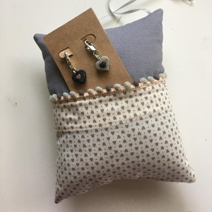 May include: A small, gray pillow with a heart-patterned fabric pocket. Two heart-shaped charms with silver clasps are attached to a brown card. The pillow has a gray top and is trimmed with pom-poms and a ribbon.
