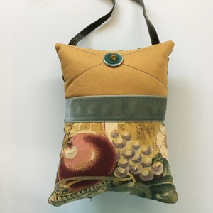May include: A handmade shoulder bag with a mustard-yellow top and a floral patterned bottom. The bag has a teal velvet band and strap. A teal button is on the front. The floral pattern includes grapes and apples. The bag is approximately 25 cm long.