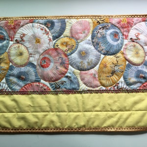 May include: A yellow quilted placemat with a border of gold satin ribbon. The top of the placemat features a colorful fabric panel with a repeating pattern of Japanese umbrellas.
