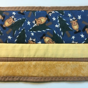 May include: A quilted placemat with a festive design. The top section features a blue background with reindeer, trees, and stars. Below are yellow and brown fabric stripes, all framed by a brown border with decorative stitching.