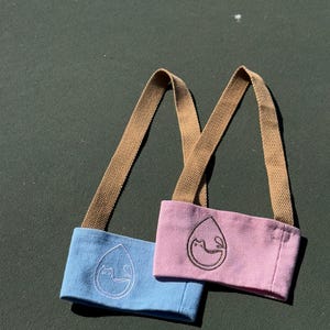 May include: Two fabric coffee cup holders with brown straps. One is light blue with a white cat design and the other is pink with a white cat design.