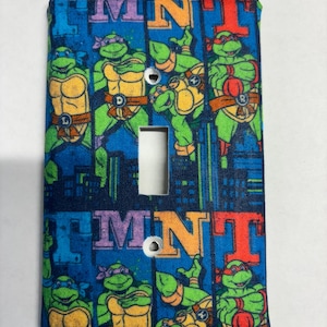 May include: A light switch cover featuring a Teenage Mutant Ninja Turtles design. The cover is blue with a repeating pattern of the four turtles, the letters "TMNT", and a cityscape.