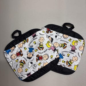 May include: Two square pot holders featuring a light gray fabric with a repeating pattern of Peanuts characters including Charlie Brown, Snoopy, Woodstock, Lucy, Linus, and Sally. Each pot holder has a solid black fabric border and a black hanging loop.