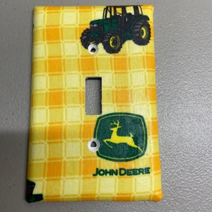 May include: A yellow and orange checkered light switch cover with a green John Deere tractor and logo. The tractor has a black cab and black wheels.