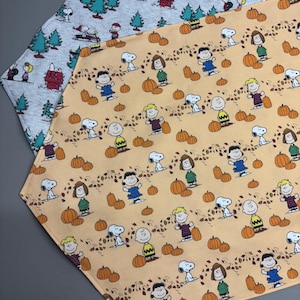 May include: Two fabric table runners. One is light gray with a pattern of Snoopy, Charlie Brown, and other Peanuts characters with Christmas trees. The other is peach-colored with Snoopy, Charlie Brown, and other Peanuts characters with pumpkins.