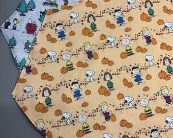 May include: Two fabric table runners. One is light gray with a pattern of Snoopy, Charlie Brown, and other Peanuts characters with Christmas trees. The other is peach-colored with Snoopy, Charlie Brown, and other Peanuts characters with pumpkins.