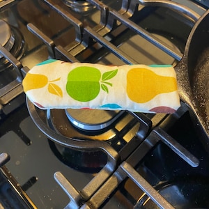 May include: A colorful pot holder with a pattern of pears and apples. The pot holder is on a black stovetop.