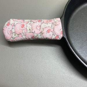 May include: A black cast iron skillet with a pink and white fabric handle cover featuring a pattern of cartoon pigs. The handle cover is designed to protect hands from heat.