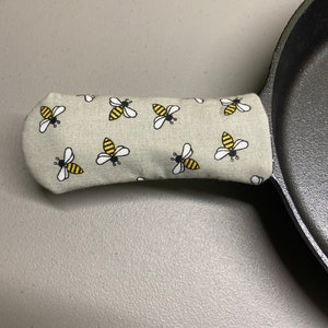 May include: A gray cast iron skillet with a removable handle. The handle is covered in a gray fabric with a repeating pattern of black and yellow bees.
