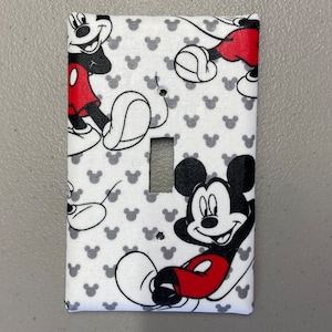 May include: A white light switch cover with a Mickey Mouse pattern. The cover features Mickey Mouse in various poses, with a red and black color scheme. The background is white with gray Mickey Mouse ears.