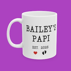 Puede incluir: Taza de cerámica blanca con el texto "BAILEY'S PAPI EST. 2025" en negro. Un corazón rojo y dos huellas de pies de bebé negras están debajo del texto. La taza tiene un asa blanca y está sobre un fondo morado.