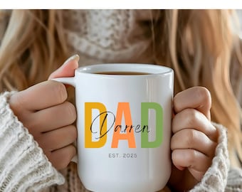 May include: White ceramic mug with the word "DAD" in yellow, orange, and green block letters. The name "Darren" is written in a cursive font, with "EST. 2025" below. The mug is filled with a dark liquid.