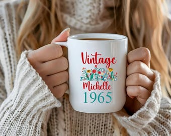 May include: White ceramic mug featuring the word "Vintage" in red script, a floral design, and the name "Michelle" and the year "1965" in teal. The mug is held by someone wearing a cream-coloured jumper.
