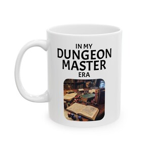 May include: White ceramic mug with black text that reads "In My Dungeon Master Era". The mug features a photo of a tabletop gaming scene with dice, books, and a wooden table.