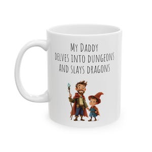 Op de afbeelding: Een witte keramische mok met een zwart-witte illustratie van een vader en zoon die verkleed zijn als fantasyfiguren. De tekst op de mok luidt "My Daddy Delves Into Dungeons And Slays Dragons."