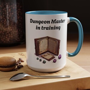 Puede incluir: Taza de cerámica blanca con interior y asa turquesa. La taza presenta el texto "Dungeon Master in training" sobre una ilustración de un libro abierto y dados. La taza está sobre una superficie de madera.