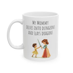 Op de afbeelding: Een witte keramische mok met een zwart-witte illustratie van een moeder en dochter die zwaarden vasthouden. De tekst op de mok luidt "My Mommy Delves Into Dungeons And Slays Dragons."
