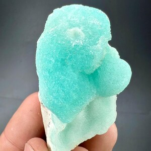 Pu&ograve; includere: Un vibrante esemplare di minerale blu chiaro con una superficie strutturata e cristallina. Il minerale ha una forma arrotondata e organica e poggia su una base bianca. Lo sfondo &egrave; grigio scuro.