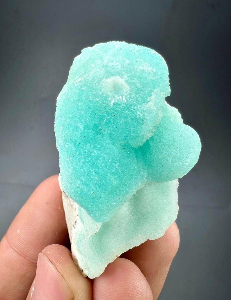 Pu&ograve; includere: Un vibrante esemplare di minerale blu chiaro con una superficie strutturata e cristallina. Il minerale ha una forma arrotondata e organica e poggia su una base bianca. Lo sfondo &egrave; grigio scuro.