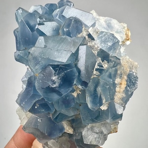 May include: A cluster of translucent blue celestite crystals with sharp, geometric facets and edges. The mineral specimen features varying shades of light to deep blue, with some areas showing clear, colorless crystalline structures at the base.