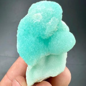 Pu&ograve; includere: Un vivace campione di minerale turchese chiaro con una superficie strutturata e cristallina. Il minerale ha una forma arrotondata e organica ed &egrave; tenuto contro uno sfondo scuro. Il colore &egrave; una tonalit&agrave; brillante e accattivante di blu-verde.