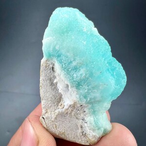 Pu&ograve; includere: Primo piano di un campione minerale con una vibrante struttura cristallina color turchese su una base ruvida grigia. La superficie del minerale ha un aspetto strutturato, con aree bianche e grigio scuro visibili.