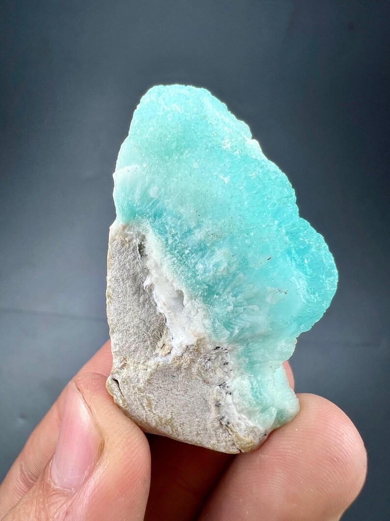 Pu&ograve; includere: Primo piano di un campione minerale con una vibrante struttura cristallina color turchese su una base ruvida grigia. La superficie del minerale ha un aspetto strutturato, con aree bianche e grigio scuro visibili.