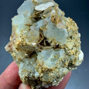 May include: A natural aquamarine crystal cluster. The specimen displays translucent, pale blue-green aquamarine crystals set in a matrix of beige and brown minerals. The rough texture and natural form are prominent.