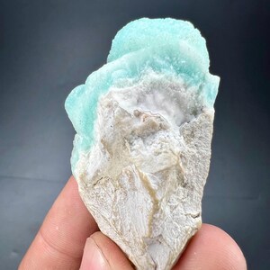 Pu&ograve; includere: Un campione di minerale naturale con una formazione cristallina azzurra su una matrice ruvida bianco sporco. Il minerale ha un aspetto strutturato, con un contrasto tra la superficie liscia blu e la base beige ruvida. Il campione &egrave; tenuto in una mano.