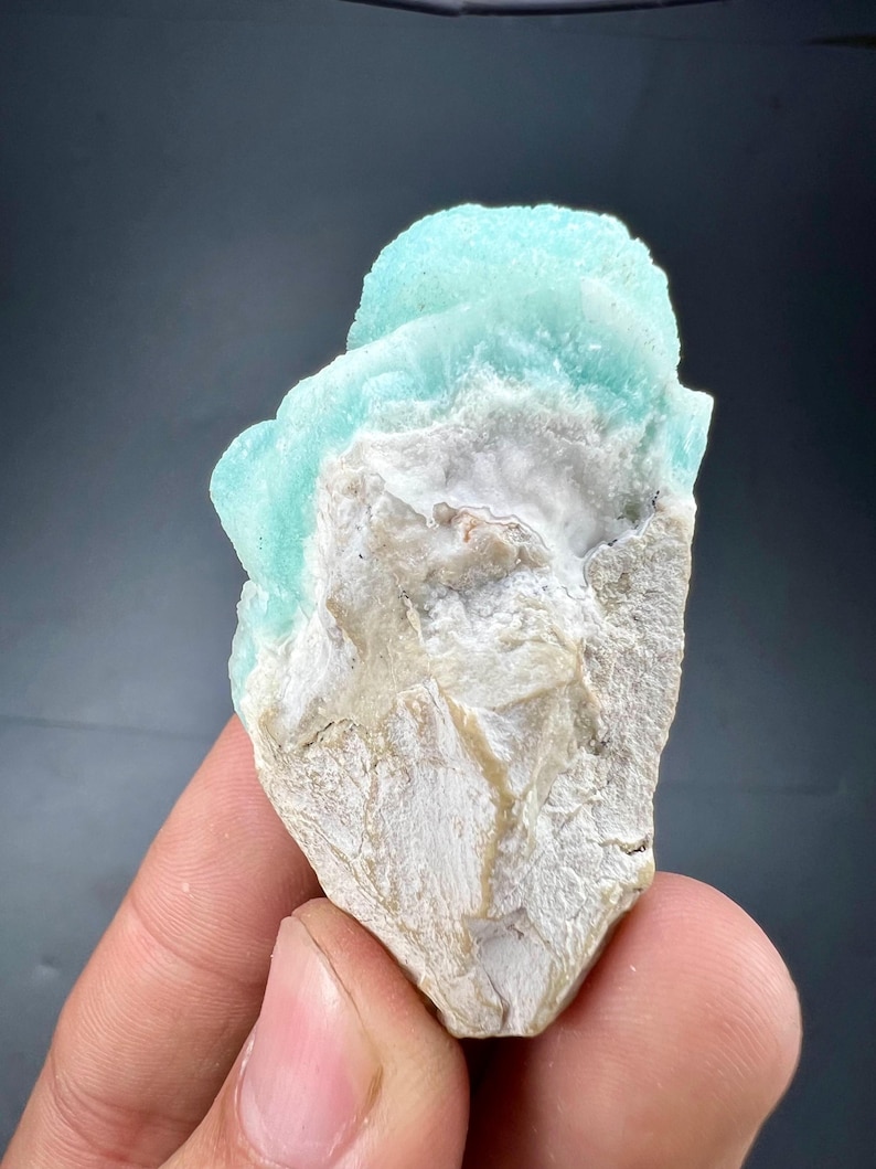 Pu&ograve; includere: Un campione di minerale naturale con una formazione cristallina azzurra su una matrice ruvida bianco sporco. Il minerale ha un aspetto strutturato, con un contrasto tra la superficie liscia blu e la base beige ruvida. Il campione &egrave; tenuto in una mano.