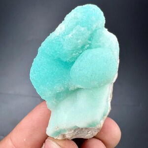 Pu&ograve; includere: Un vibrante esemplare minerale blu chiaro con una superficie strutturata e cristallina. Il minerale ha una forma arrotondata e organica e poggia su una base ruvida bianco sporco. L'esemplare &egrave; tenuto contro uno sfondo scuro.