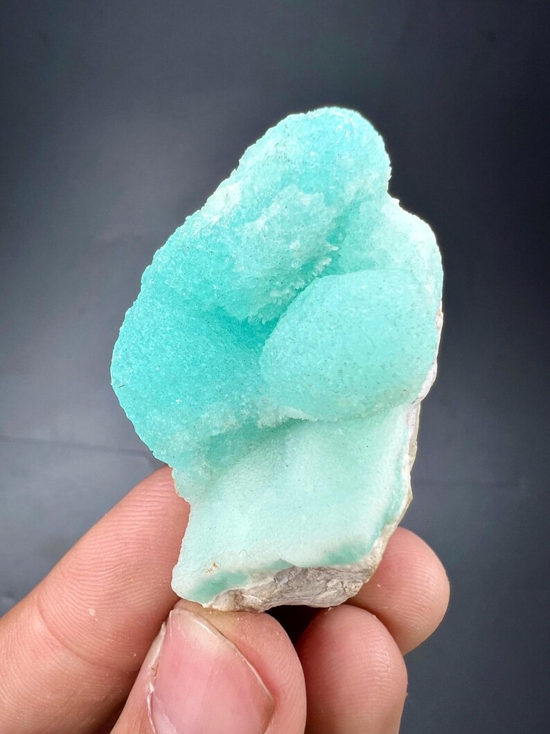 Pu&ograve; includere: Un vibrante esemplare minerale blu chiaro con una superficie strutturata e cristallina. Il minerale ha una forma arrotondata e organica e poggia su una base ruvida bianco sporco. L'esemplare &egrave; tenuto contro uno sfondo scuro.