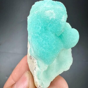 Pu&ograve; includere: Un vibrante esemplare minerale blu chiaro con una superficie strutturata e cristallina. Il minerale ha una forma arrotondata e organica e poggia su una base di colore chiaro. Lo sfondo &egrave; grigio scuro.