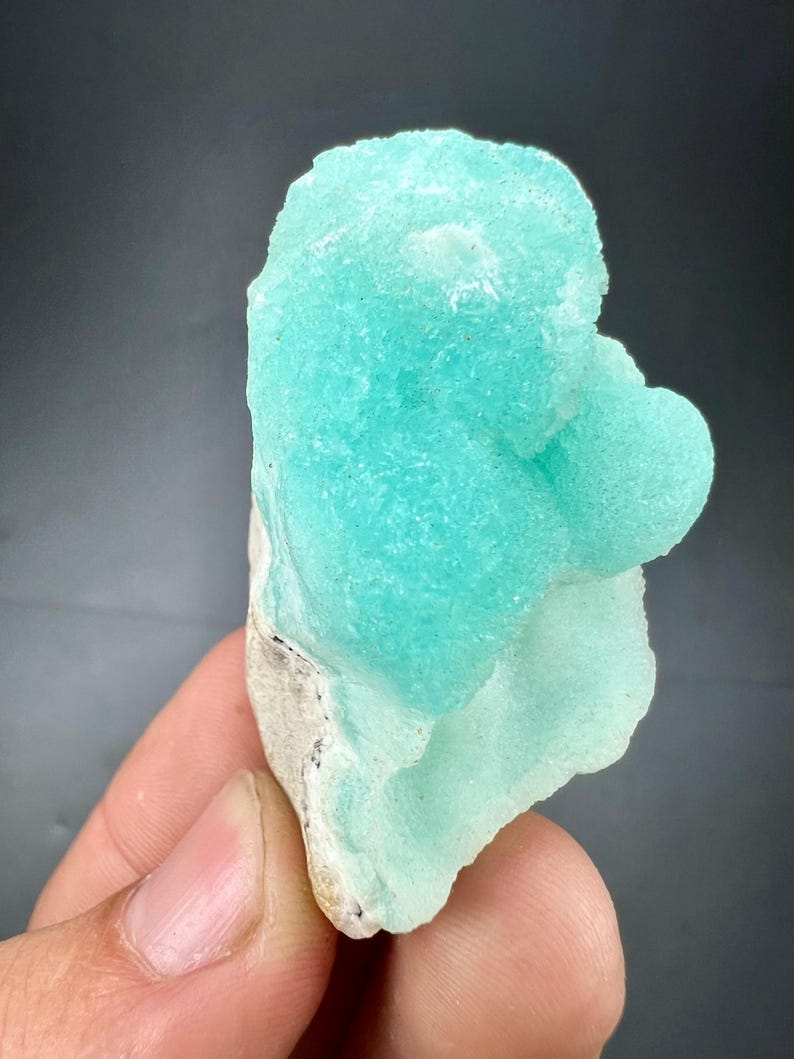 Pu&ograve; includere: Un vibrante esemplare minerale blu chiaro con una superficie strutturata e cristallina. Il minerale ha una forma arrotondata e organica e poggia su una base di colore chiaro. Lo sfondo &egrave; grigio scuro.