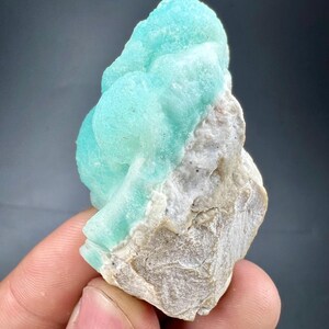 Pu&ograve; includere: Un vibrante campione minerale azzurro con una superficie strutturata e cristallina. Il minerale &egrave; posto su una base di roccia ruvida color beige. Il campione &egrave; tenuto in mano su uno sfondo grigio scuro.