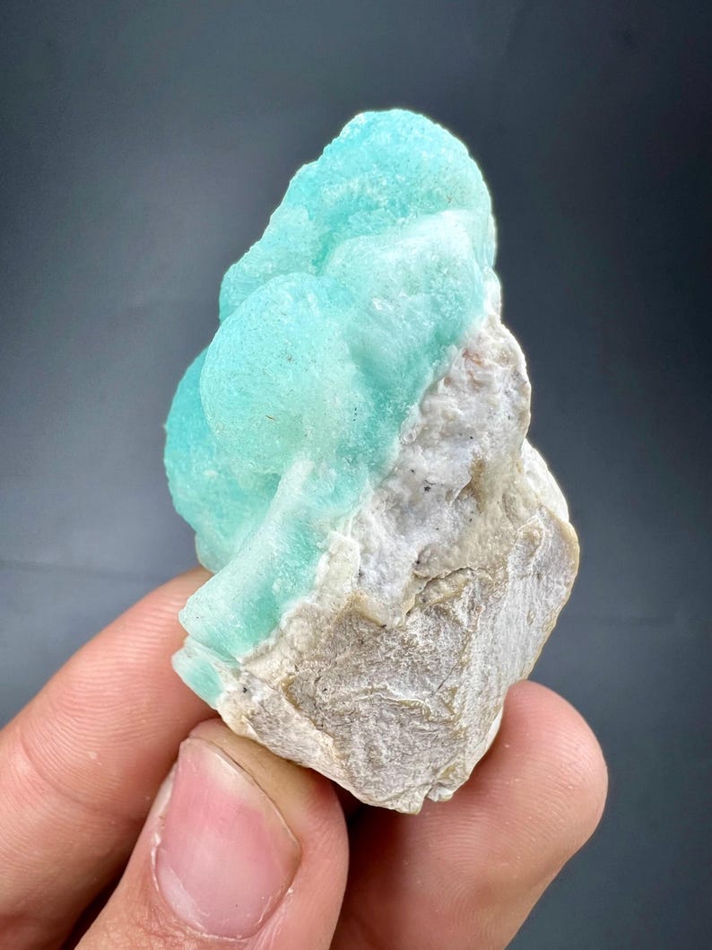 Pu&ograve; includere: Un vibrante campione minerale azzurro con una superficie strutturata e cristallina. Il minerale &egrave; posto su una base di roccia ruvida color beige. Il campione &egrave; tenuto in mano su uno sfondo grigio scuro.