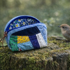May include: A small, open, patchwork fabric pouch with a blue zipper and a brown leather pull. The pouch is made of various fabric pieces in blue, yellow, green, and white. The interior lining features a white daisy pattern on a blue background.