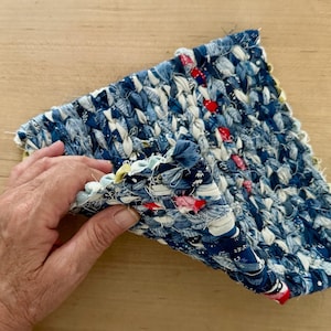 May include: A handmade, woven textile in shades of blue and white, with accents of red and pink. The fabric is held up, revealing the texture and construction of the piece. The textile appears to be a small rug or mat.