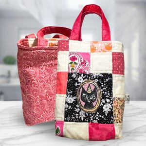May include: Two small tote bags. The bag on the right is patchwork with pink, white, and black squares. It features a black cat with a unicorn horn. The bag on the left is a pink floral pattern.
