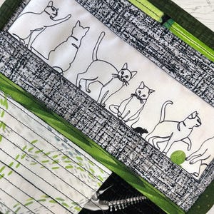 Puede incluir: Un artículo de tela acolchada con un panel blanco con dibujos lineales negros de gatos en varias poses. El panel está bordeado por tela estampada gris y verde. Una pequeña bola verde está cerca de uno de los gatos.