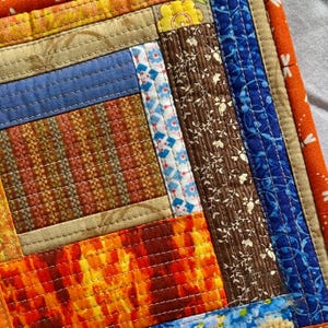 May include: Close-up of a handmade patchwork quilt with a variety of fabrics. The quilt features squares and rectangles in shades of orange, blue, brown, and yellow. The quilt is stitched with white thread.