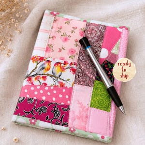 May include: A patchwork journal cover in shades of pink, featuring floral and bird patterns. A silver pen rests on the cover. The cover is bordered with a green floral fabric. A round sticker says "ready to ship".
