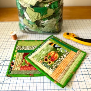 May include: Two square, quilted fabric coasters with green borders. One coaster features a pumpkin and sunflowers, the other has a red apple. A bag of green fabric scraps and a spool of thread are in the background.