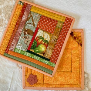 May include: Two handmade quilted pot holders. The top pot holder features a patchwork design with various fabrics in shades of orange, green, red, and yellow. The center showcases a pumpkin print. The second pot holder is orange with a floral leather tag.