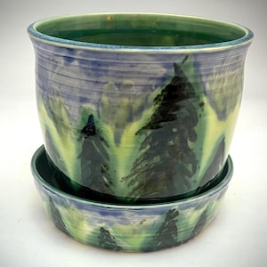 May include: A ceramic planter with a matching saucer. The planter is cylindrical with a wide opening, painted with a landscape scene of green trees and blue sky. The saucer mirrors the design. The planter is suitable for indoor or outdoor use.