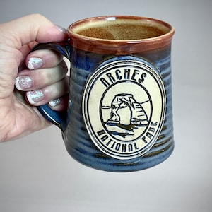 Könnte beinhalten: Eine blaue und braune Keramiktasse mit einem weißen Kreisdesign mit dem Text "Arches National Park" und einer Darstellung eines natürlichen Bogens.