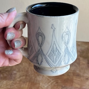 May include: Cream-colored ceramic mug with a black interior and a decorative pattern in dark blue. The mug features a handle and a pedestal base. The design includes geometric shapes and lines. The mug is held by a hand with glittery nail polish.