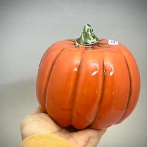 May include: A glossy orange ceramic pumpkin with a green stem. The pumpkin has vertical ridges and is held in a hand. This decorative item is perfect for autumn and Halloween.