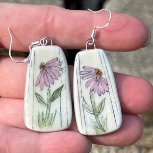 Puede incluir: Pendientes de cerámica pintados a mano con un diseño de flor rosa. Los pendientes tienen forma rectangular con fondo blanco y líneas negras verticales. El diseño floral incluye hojas y tallos verdes, con ganchos plateados.