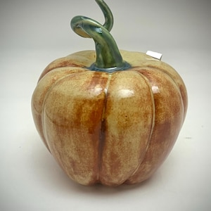 Puede incluir: Una calabaza de cerámica con acabado brillante. La calabaza es principalmente de color marrón claro con rayas verticales marrones oscuras y un tallo verde. El tallo se curva ligeramente. Decoración de otoño.